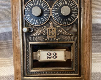 May include: A vintage-style post office box door set in a light oak wood frame. The brass-toned metal face features two circular combination dials with letters, a central eagle emblem with the letters US, and a glass window displaying the number 23.