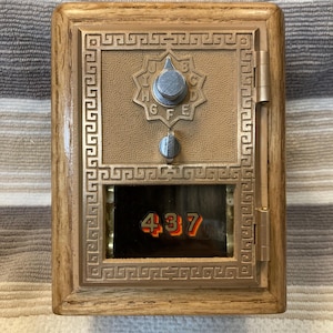 May include: A vintage-style post office box door with a wooden frame, featuring a brass-colored metal faceplate with a Greek key border. It includes a circular dial with letters A through J, a small latch, and a glass window displaying the number 437.