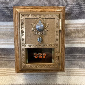 Puede incluir: Un buzón vintage con marco de madera y frente de metal color bronce. El frente de metal presenta un dial en forma de estrella con letras y una pequeña ventana que muestra el número 357 en naranja. El buzón tiene una puerta con bisagras y un borde decorativo.