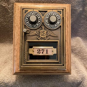Peut inclure: Une boîte aux lettres vintage en laiton et en bois avec un cadre rectangulaire. La boîte aux lettres comporte deux cadrans circulaires avec des lettres, un emblème d'aigle américain et le numéro "271" affiché sur une petite plaque. Le cadre en bois a une finition naturelle.