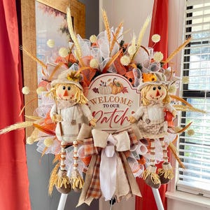 May include: A fall-themed wreath with two scarecrow figures and a sign that reads "Welcome to our Patch." The wreath features orange, white, and brown ribbons, wheat stalks, and decorative elements. The scarecrows wear hats and overalls.