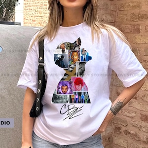 Op de afbeelding: Wit T-shirt met een silhouet gevuld met verschillende afbeeldingen en een handtekening. Het shirt wordt gecombineerd met een lichtblauwe jeans. De tekst "NERMIKASTUDIO" is zichtbaar. Korte mouwen.