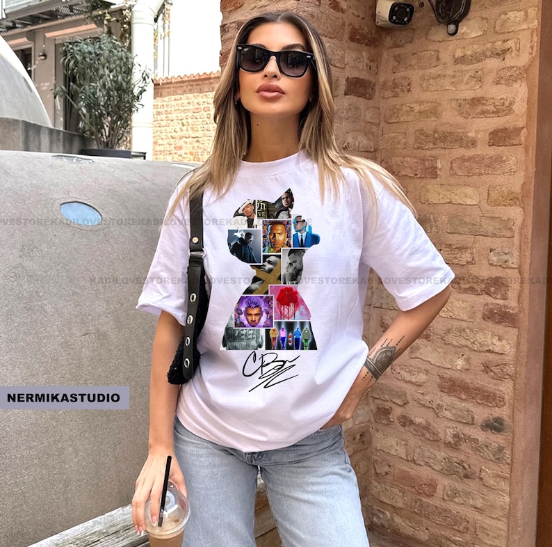 Op de afbeelding: Wit T-shirt met een silhouet gevuld met verschillende afbeeldingen en een handtekening. Het shirt wordt gecombineerd met een lichtblauwe jeans. De tekst "NERMIKASTUDIO" is zichtbaar. Korte mouwen.