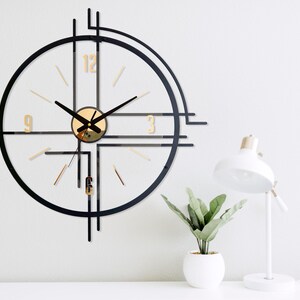 May include: A modern wall clock with a black frame and gold accents. The clock features a circular design with gold numerals and hands. A white desk lamp and a small potted plant are also visible.