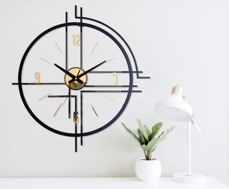 May include: A modern wall clock with a black frame and gold accents. The clock features a circular design with gold numerals and hands. A white desk lamp and a small potted plant are also visible.