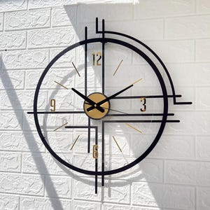 May include: A modern wall clock with a black frame and gold accents. The clock face is transparent, with gold-colored numerals at 3, 6, 9, and 12. The hands are black, and the clock is mounted on a white brick wall.