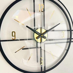 May include: A modern wall clock with a transparent face, black frame, and gold accents. The clock features gold numerals at 3, 6, 9, and 12, with gold stick markers for the other hours. The hands are black.
