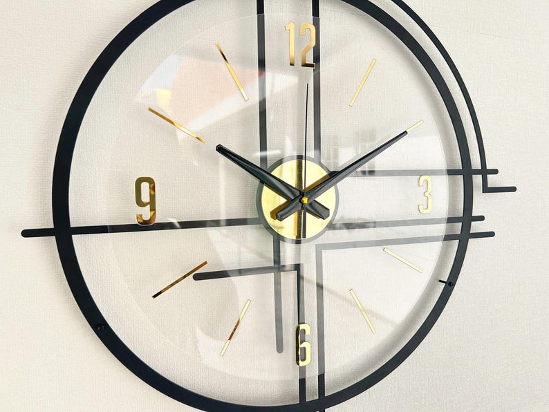 May include: A modern wall clock with a transparent face, black frame, and gold accents. The clock features gold numerals at 3, 6, 9, and 12, with gold stick markers for the other hours. The hands are black.
