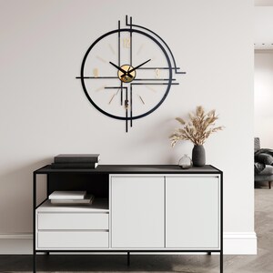 May include: A modern wall clock with a black frame and gold accents. The clock has a circular design with a clear face and gold numerals. The clock is mounted on a white wall above a black and white cabinet.