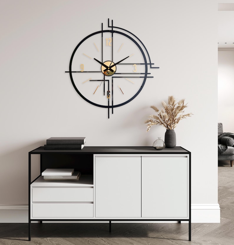 May include: A modern wall clock with a black frame and gold accents. The clock has a circular design with a clear face and gold numerals. The clock is mounted on a white wall above a black and white cabinet.