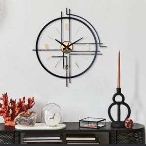 May include: A modern wall clock with a black metal frame and gold accents. The clock has a circular design with a minimalist aesthetic. The clock is mounted on a white wall, with decorative items on a black table below.