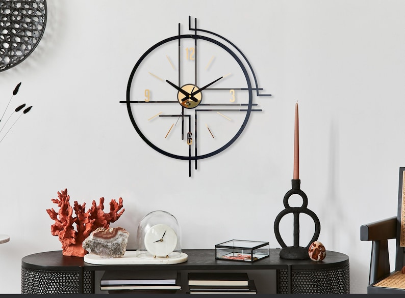 May include: A modern wall clock with a black metal frame and gold accents. The clock has a circular design with a minimalist aesthetic. The clock is mounted on a white wall, with decorative items on a black table below.