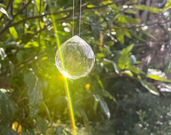 Puede incluir: Una bola de cristal transparente colgada de una rama de árbol, con la luz del sol brillando a través de ella, creando un efecto arcoíris.
