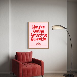 Puede incluir: Una impresi&oacute;n enmarcada rosa y blanca con el texto "You're Doing Amazing Sweetie" en rojo. La impresi&oacute;n est&aacute; colgada en una pared blanca sobre un sill&oacute;n rojo.