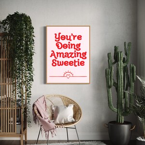 Puede incluir: Una impresi&oacute;n enmarcada en rosa y blanco con el texto "You're Doing Amazing Sweetie" en rojo. La impresi&oacute;n tambi&eacute;n presenta un coraz&oacute;n rojo y un sol rojo.