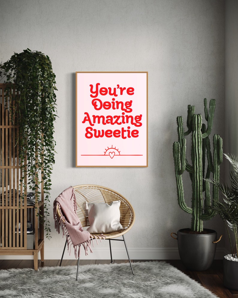 Puede incluir: Una impresi&oacute;n enmarcada en rosa y blanco con el texto "You're Doing Amazing Sweetie" en rojo. La impresi&oacute;n tambi&eacute;n presenta un coraz&oacute;n rojo y un sol rojo.