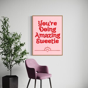 Puede incluir: Una impresi&oacute;n enmarcada rosa con el texto "You're Doing Amazing Sweetie" en letras rojas. Un peque&ntilde;o sol con un coraz&oacute;n dentro est&aacute; debajo del texto.