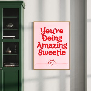 Puede incluir: Un p&oacute;ster motivacional rosa y rojo con el texto "You're Doing Amazing Sweetie" y un peque&ntilde;o sol con un coraz&oacute;n en el centro.