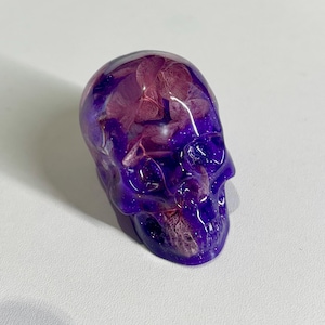 May include: A purple resin skull with embedded dried flowers. The skull has a glossy finish and is a deep shade of purple, with hints of pink and white from the flowers. The skull is a decorative item.