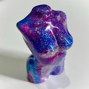 May include: A resin torso sculpture, adorned with blue, purple, and silver glitter. The form is a stylized representation of the upper body. The sculpture is a decorative item, suitable for display. The glitter adds a shimmering effect to the piece.