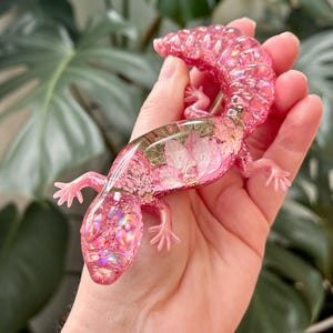 May include: A translucent pink resin gecko figurine featuring embedded dried pink flowers and iridescent glitter throughout its body. The lizard has a curved tail, four small legs, and a smooth, glossy finish with a shimmering, reflective appearance.