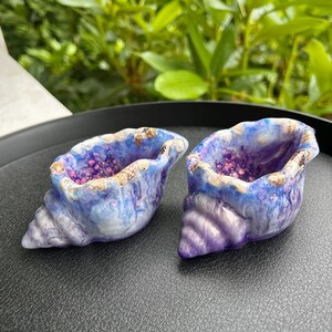 May include: Two shell-shaped trinket dishes featuring a marbled blue and purple finish with embedded glitter accents. The edges of the dishes are textured with small, realistic shell details, creating a decorative container for small items.