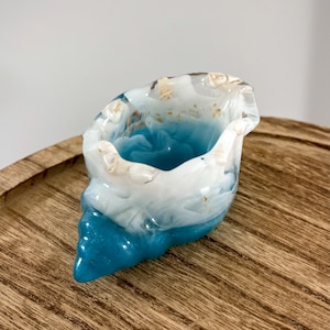 Peut inclure: Un récipient décoratif en forme de coquillage avec un intérieur bleu vif et des accents blancs et dorés. La pièce en résine a une finition brillante et est posée sur un plateau en bois. Le récipient mesure environ 10 cm de long.