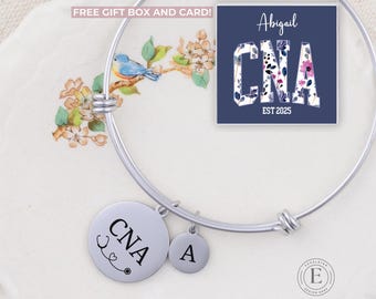 May include: A silver charm bracelet with a charm that says "CNA" with a stethoscope and heart. There is also a charm with the letter "A". The bracelet is on a white surface with a blue card that says "Abigail CNA EST 2025" with floral designs.