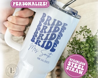 May include: White travel mug with a handle, featuring the word "BRIDE" in a retro blue font. The mug is personalized with "Mrs. Smith" and the date "08.16.2025". Includes a stainless steel straw. The mug is designed for a wedding.
