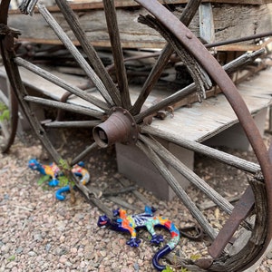 May include: An antique wooden wagon wheel with a rusted metal rim. Two colorful ceramic lizard figurines with blue, orange, and green details rest on the ground near the wheel. The scene is set outdoors.