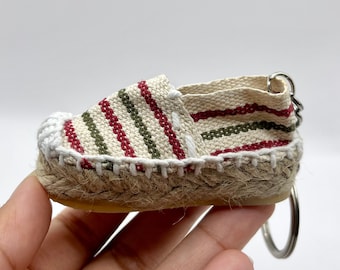 May include: A small, striped espadrille shoe keychain with a red, green, and white striped pattern. The shoe is made of a woven fabric and has a rope sole.