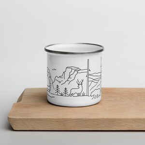 May include: White enamel camping mug with a black line drawing of a mountain landscape, including trees, a deer, and the word "Yosemite". The mug has a silver rim and is sitting on a wooden cutting board.