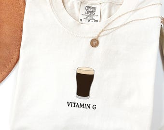 May include: Ivory t-shirt featuring a graphic of a dark stout beer and the text "VITAMIN G". A gold necklace and a brown hat are next to the shirt. The shirt is made by Comfort Colors.