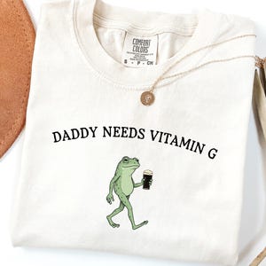 K&ouml;nnte beinhalten: Elfenbeinfarbenes Sweatshirt mit dem Text "DADDY NEEDS VITAMIN G" und einer Cartoon-Fr&ouml;schin, die ein dunkles Getr&auml;nk h&auml;lt. Das Shirt hat einen Rundhalsausschnitt und ist mit "COMFORT COLORS" gekennzeichnet. Eine goldene Halskette und ein brauner Hut sind ebenfalls zu sehen.
