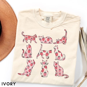 May include: Ivory-colored t-shirt featuring a playful design of cats with strawberry patterns. The cats are in various poses, with a red and pink color scheme. The shirt has a crew neck and a tag that says "Comfort Colors". The word "IVORY" is printed at the bottom.