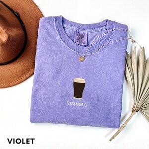 Pu&ograve; includere: Una maglietta Comfort Colors viola con una grafica di una pinta di stout e la scritta "Vitamin G". Una collana d'oro con un ciondolo &egrave; appoggiata sulla maglietta. La parola "VIOLET" &egrave; stampata in basso.