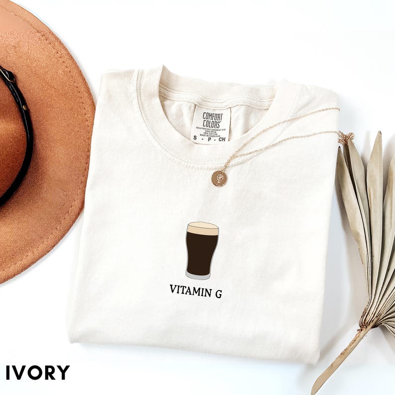 Pu&ograve; includere: T-shirt color avorio con una grafica di una birra scura e il testo "VITAMIN G". Una collana d'oro e un cappello marrone sono accanto alla maglietta. La maglietta &egrave; di Comfort Colors.