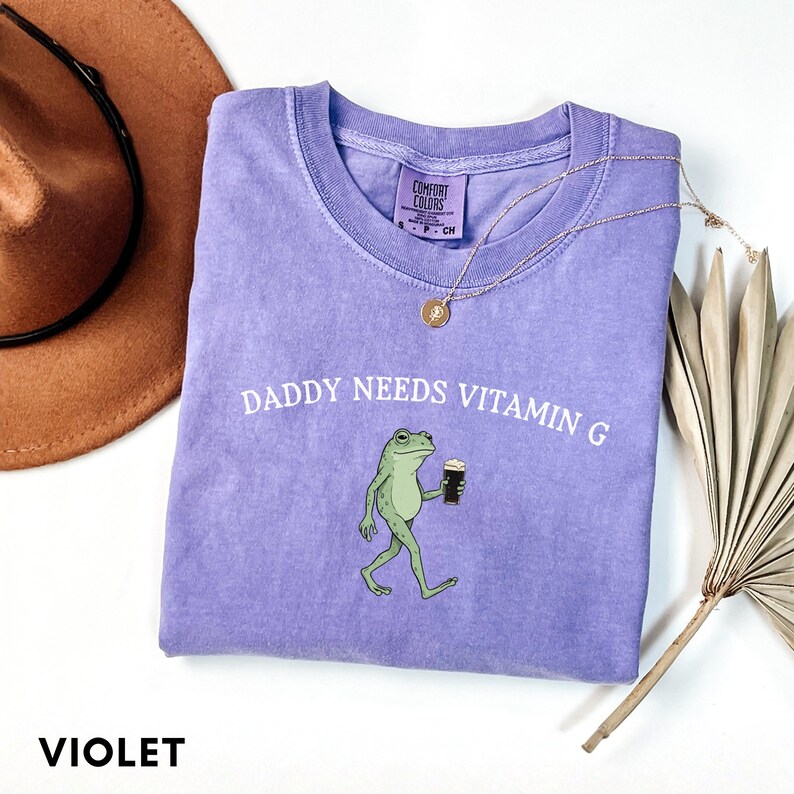 K&ouml;nnte beinhalten: Violettes T-Shirt mit dem Schriftzug "DADDY NEEDS VITAMIN G" und einem Cartoon-Frosch, der ein dunkles Getr&auml;nk h&auml;lt. Das Shirt hat einen Rundhalsausschnitt und ein kleines Etikett. Eine goldene Halskette und ein brauner Hut sind ebenfalls sichtbar.