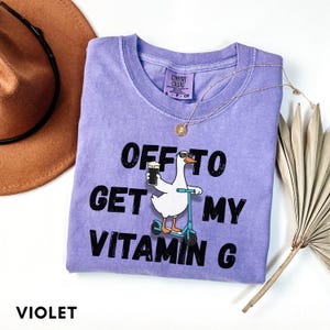 Puede incluir: Camiseta violeta Comfort Colors con un gr&aacute;fico de un ganso con gafas de sol montado en un scooter y sosteniendo una cerveza. El texto de la camiseta dice "OFF TO GET MY VITAMIN G". Tambi&eacute;n se ve un collar dorado y un sombrero marr&oacute;n.