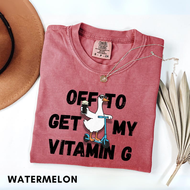 Puede incluir: Una camiseta roja con la frase "OFF TO GET MY VITAMIN G" y un dibujo de un ganso en un scooter sosteniendo una cerveza. La camiseta tiene cuello redondo y una peque&ntilde;a etiqueta. Tambi&eacute;n se ven un collar plateado y un sombrero.