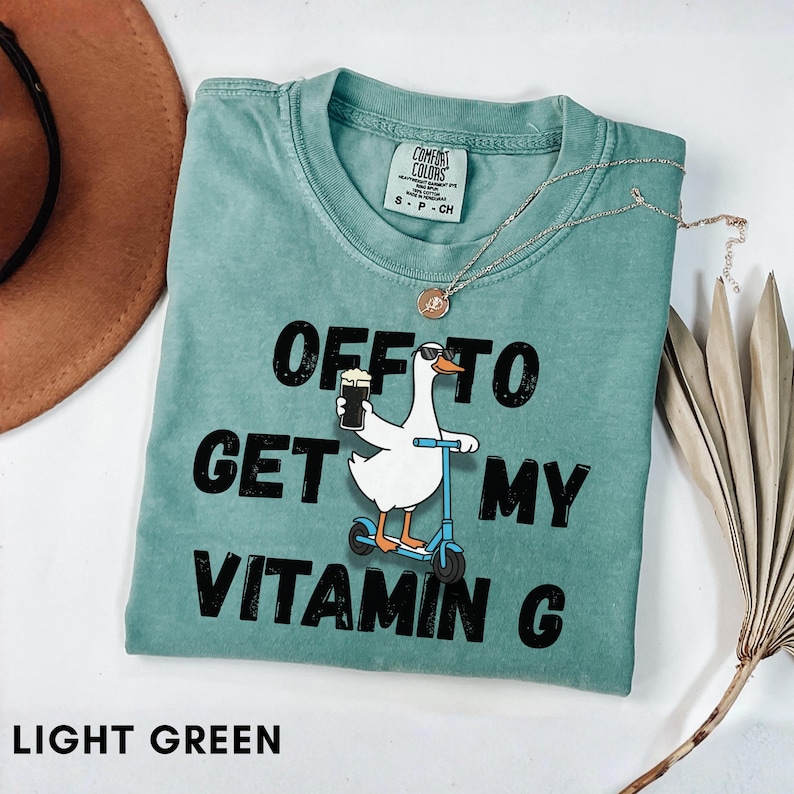 Puede incluir: Camiseta verde claro Comfort Colors con el texto "OFF TO GET MY VITAMIN C". El dise&ntilde;o presenta un ganso en un scooter sosteniendo una cerveza. Tambi&eacute;n se ven un collar dorado y un sombrero marr&oacute;n.
