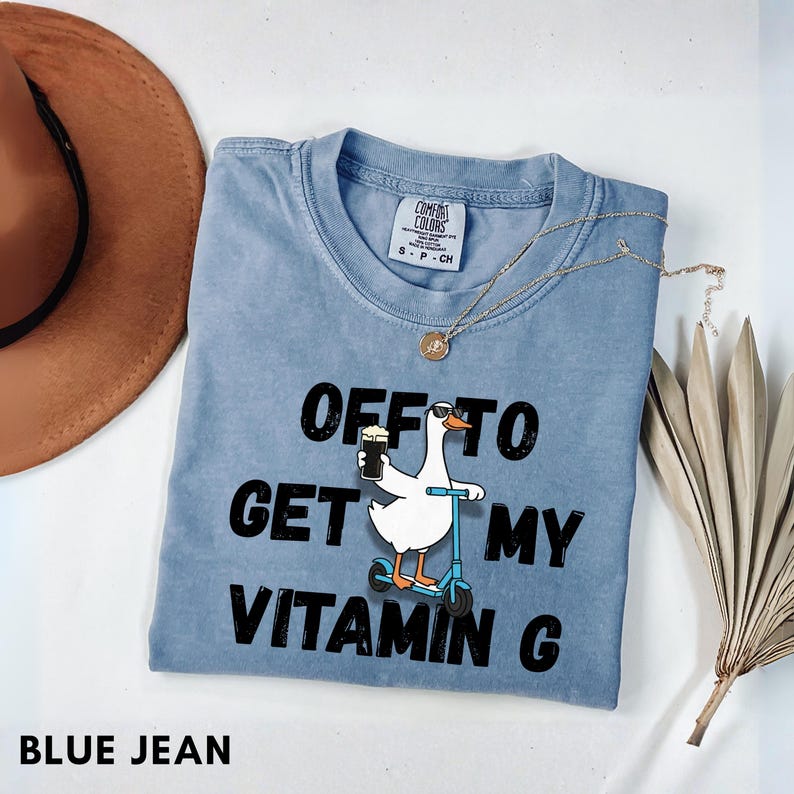 Puede incluir: Camiseta Comfort Colors azul vaquero con la frase "OFF TO GET MY VITAMIN G" y un dibujo de un ganso en un patinete con una cerveza. Un collar dorado y un sombrero marr&oacute;n tambi&eacute;n est&aacute;n en la imagen.