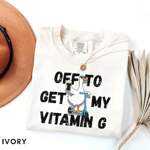 Puede incluir: Camiseta Comfort Colors color marfil con el texto "OFF TO GET MY VITAMIN G". El dise&ntilde;o presenta un ganso de dibujos animados en un scooter con una bebida. Tambi&eacute;n se ve un collar dorado y un sombrero marr&oacute;n.
