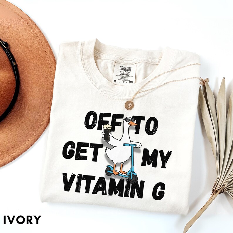 Puede incluir: Camiseta Comfort Colors color marfil con el texto "OFF TO GET MY VITAMIN G". El dise&ntilde;o presenta un ganso de dibujos animados en un scooter con una bebida. Tambi&eacute;n se ve un collar dorado y un sombrero marr&oacute;n.