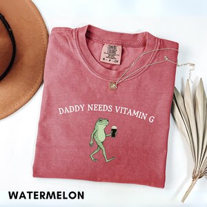 K&ouml;nnte beinhalten: Ein rotes T-Shirt mit dem Aufdruck "DADDY NEEDS VITAMIN G" und einer Cartoon-Froschfigur, die ein Getr&auml;nk h&auml;lt. Das T-Shirt hat einen Rundhalsausschnitt und ein kleines Etikett. Eine silberne Halskette und ein brauner Hut sind ebenfalls zu sehen.