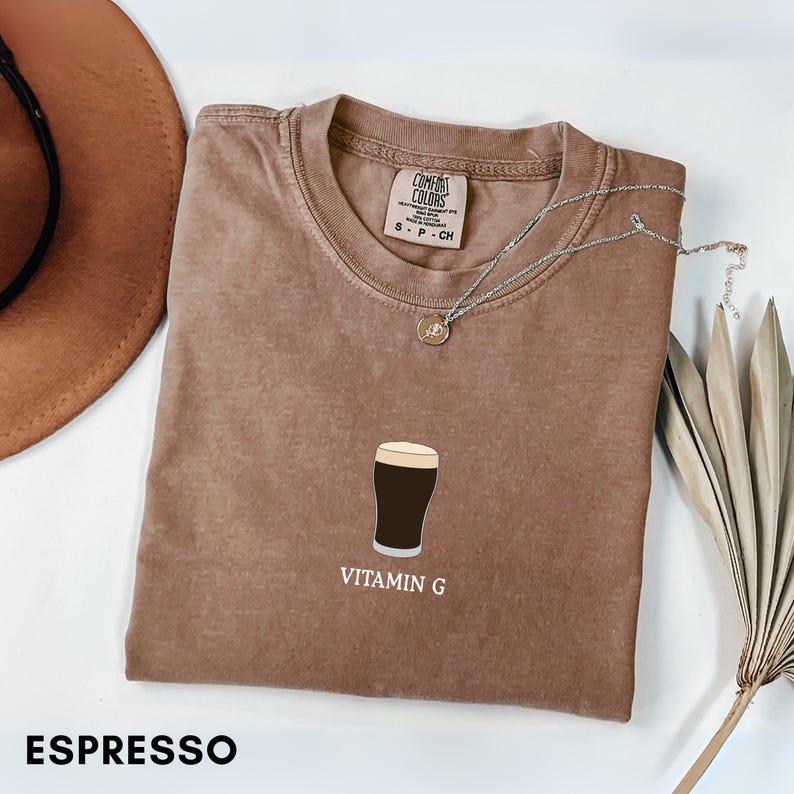 Pu&ograve; includere: T-shirt color espresso con grafica di una birra scura e la scritta "VITAMIN G". La maglietta Comfort Colors ha girocollo e maniche corte. Una collana d'argento con pendente &egrave; drappeggiata sulla maglietta.
