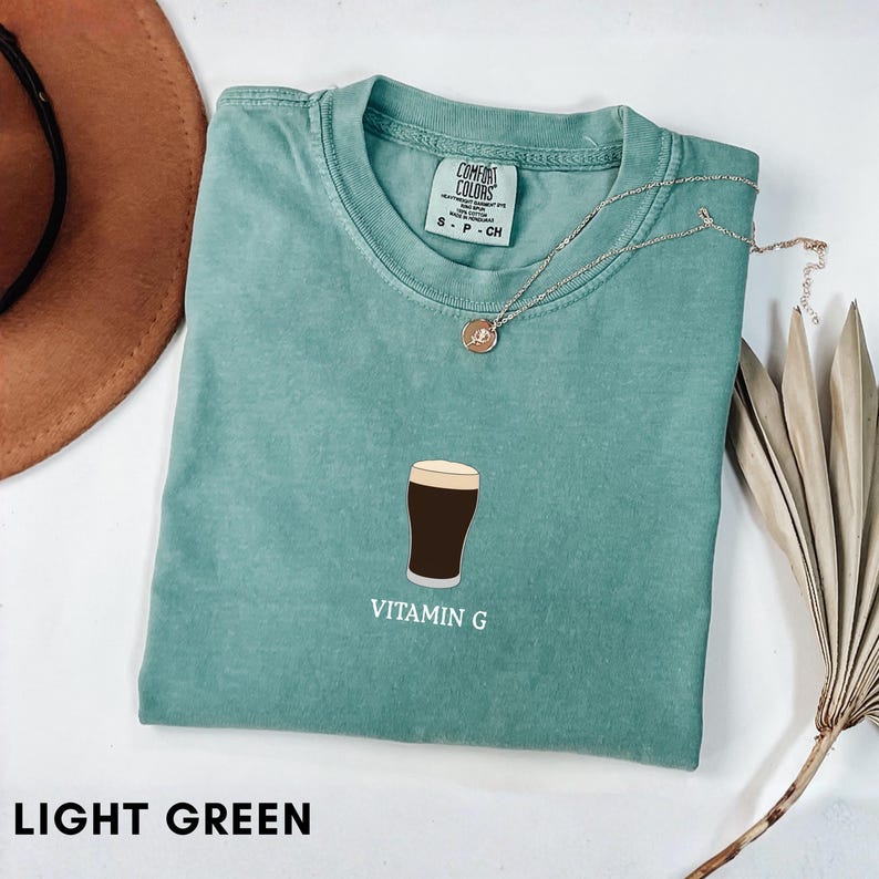 Pu&ograve; includere: T-shirt verde chiaro con una grafica di una pinta di birra e la scritta "Vitamin G". La maglietta &egrave; piegata e ha un'etichetta Comfort Colors. Sono visibili anche una collana d'argento e un cappello marrone.