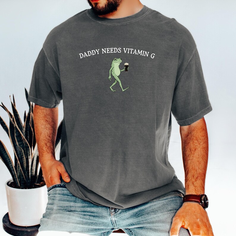 K&ouml;nnte beinhalten: Dunkelgraues T-Shirt mit dem Text "DADDY NEEDS VITAMIN G" &uuml;ber einer Cartoon-Fr&ouml;schin, die ein dunkles Getr&auml;nk h&auml;lt. Der Frosch ist gr&uuml;n und der Text ist wei&szlig;. Das Shirt hat kurze &Auml;rmel.