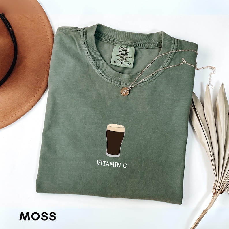 Pu&ograve; includere: Una maglietta verde muschio con una grafica di una birra scura in un bicchiere, con la scritta "Vitamin G". Visibili una collana d'oro e un cappello marrone. La maglietta &egrave; in materiale Comfort Colors.