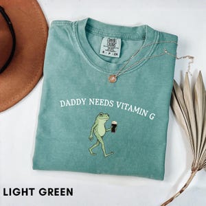 K&ouml;nnte beinhalten: Hellgr&uuml;nes T-Shirt mit dem Text "DADDY NEEDS VITAMIN G" und einer Cartoon-Fr&ouml;schin, die ein Getr&auml;nk h&auml;lt. Das Shirt hat einen Rundhalsausschnitt und kurze &Auml;rmel. Eine silberne Halskette mit Anh&auml;nger liegt auf dem Shirt.